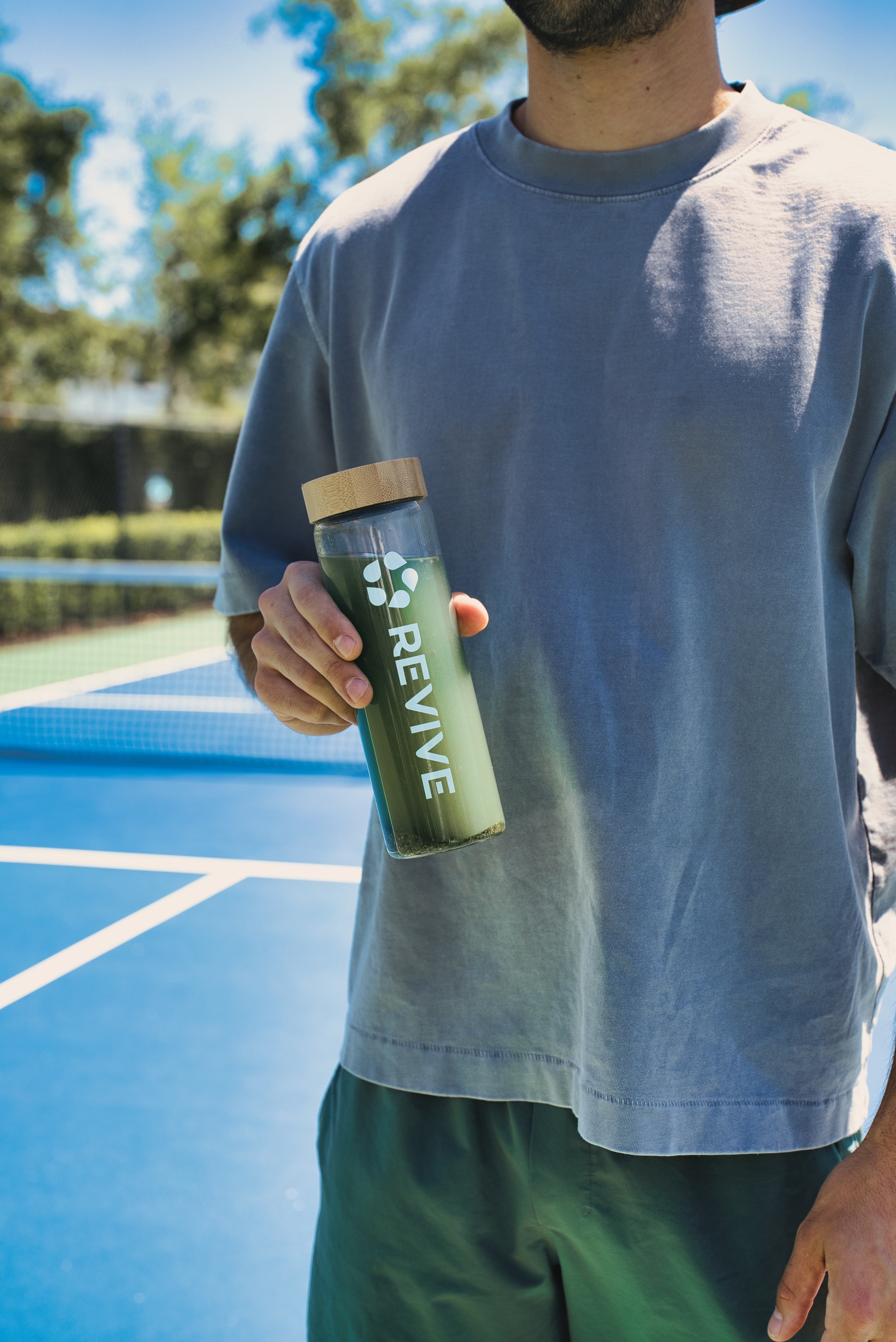 Man standing on an outdoor tennis court holding a glass bottle with green liquid and a cork lid, featuring the Revive logo printed vertically on the bottle