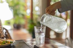 Close-up of a hand pouring cold water from a glass pitcher into a glass, with ice and condensation visible. Refreshing drink on a wooden table with salad in the background.