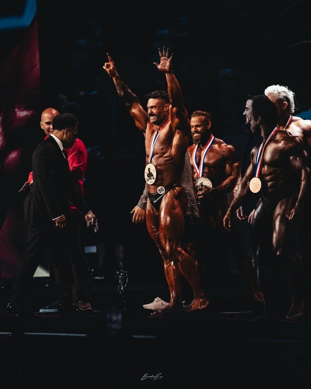 Group of male bodybuilders on stage during an award ceremony, with one competitor in the center raising both arms in celebration while wearing a medal and competition number 58